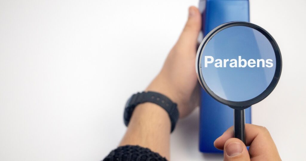 Chemical components on the shampoo label: parabens . A hand holds a blue jar and a magnifier, where the harmful ingredients of a detergent are written in close up.