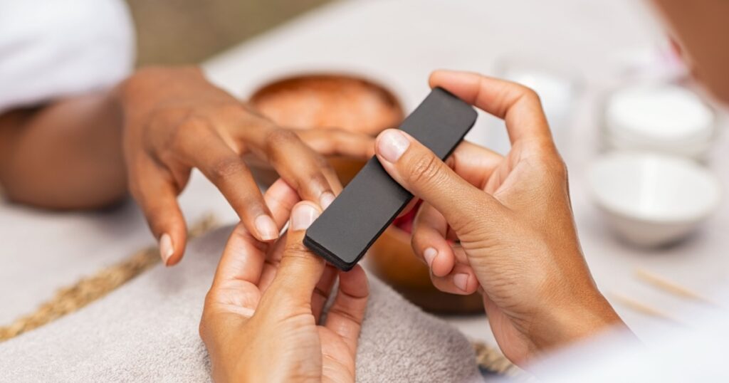 Close up of african hands of a qualified manicurist filing the nails of a young woman. Hands during manicure care session. Detail of a girl in a nail salon receiving manicure.