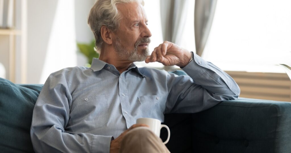 Pensive elderly 60s man sit relax on couch in living room drinking tea look in window distance thinking, thoughtful mature 50s husband rest on sofa at home feel sad pondering considering future
