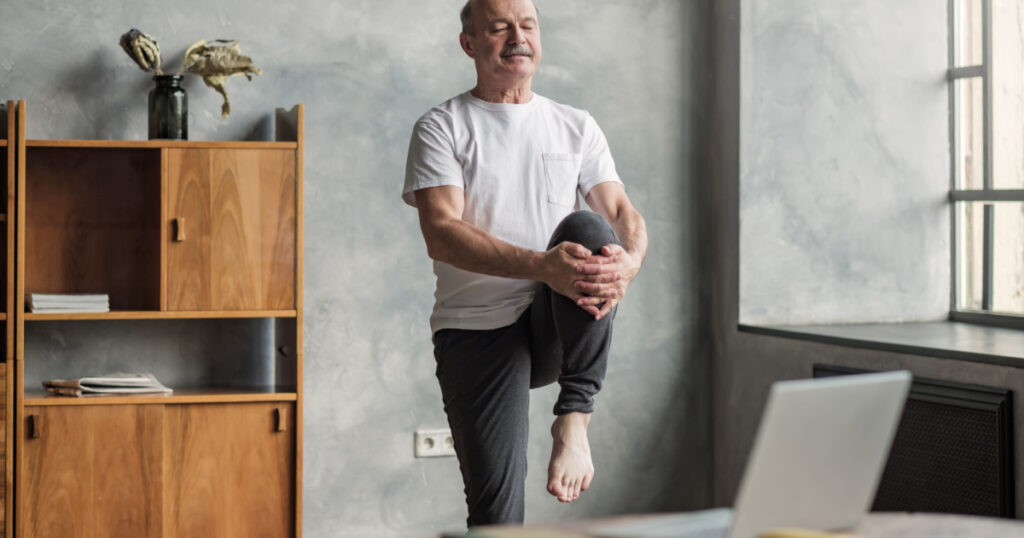 Senior handsome sportman doing stretching and balancing yoga exercise at home using online lesson on notebook. How to stay healthy on quarantine concept.