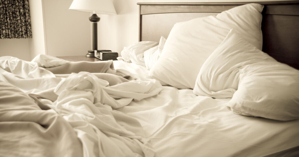 An unmade bed with white linens and a lamp in the corner.