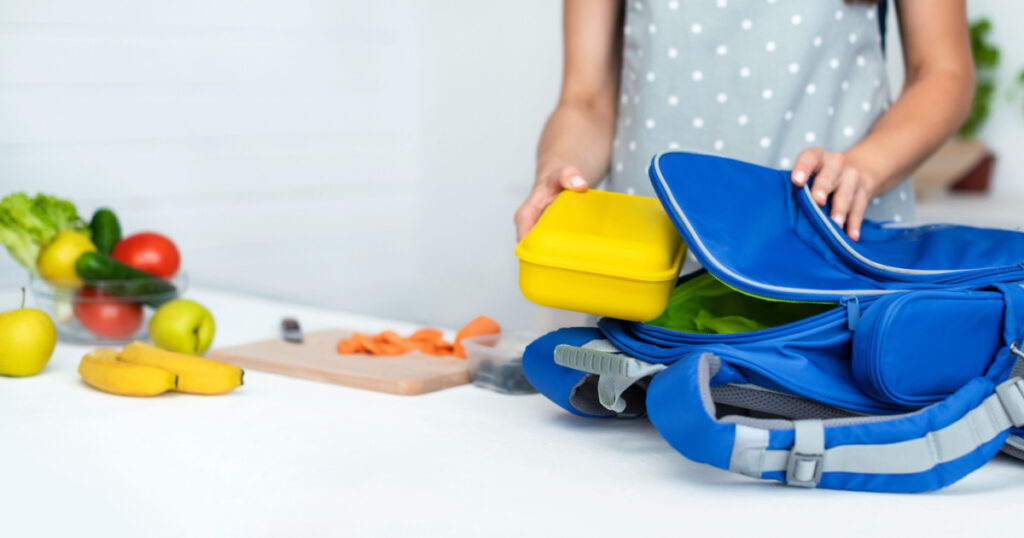 Caring mother puts yellow plastic lunch box to her son in a school backpack. School food or lunch, concept image. Selective focus, close-up.