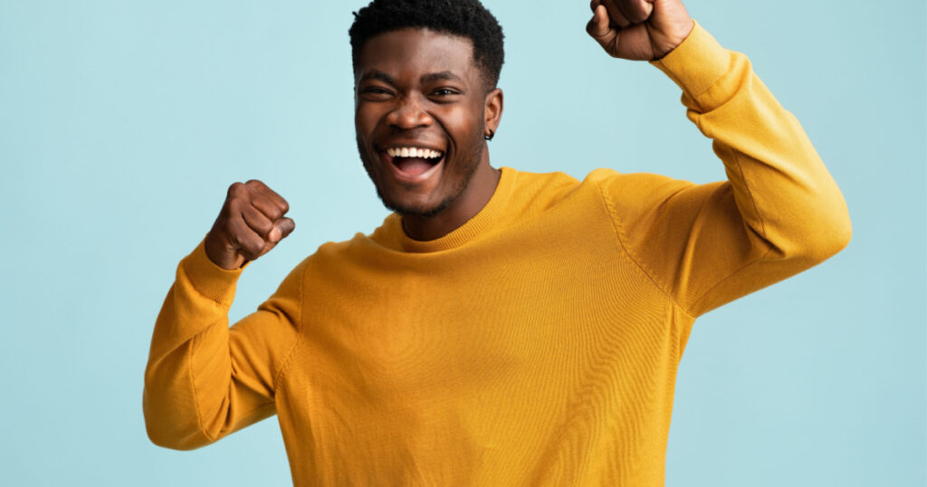 Emotional young black guy in casual raising fists up on blue studio background, celebrating success, happy handsome african american millennial man sharing good emotions, copy space