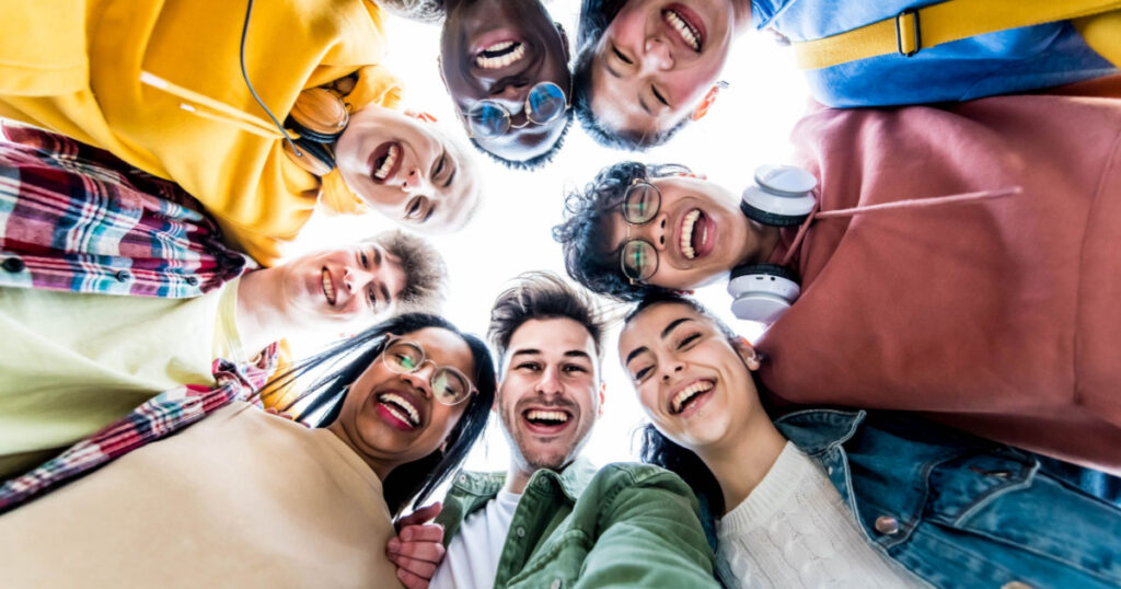 Multiracial group of young people standing in circle smiling at camera - International teamwork students hugging together - Human resources and youth culture concept