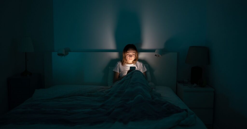 Sleepy exhausted woman lying in bed using smartphone, can not sleep. Insomnia, addiction concept. Sad girl bored in bed scrolling through social networks on mobile phone late at night in dark bedroom.