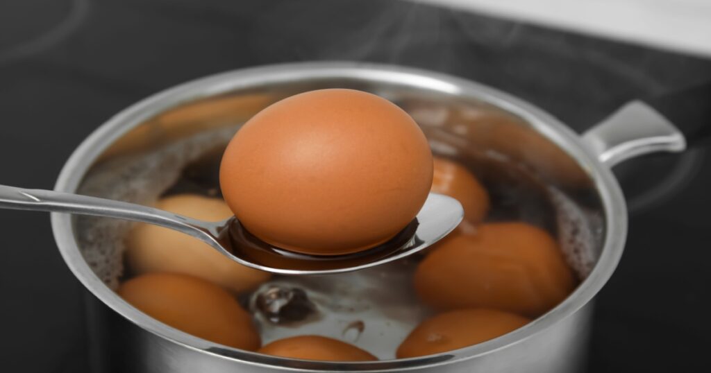 Spoon with boiled egg above saucepan on electric stove, closeup
