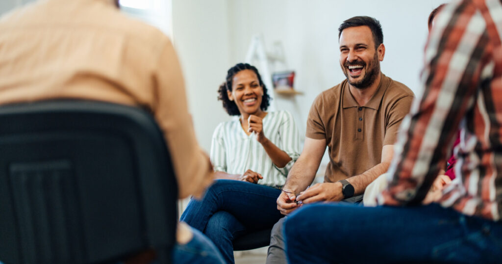 Focus on the smiling man, talking with people of all ages, during the group therapy.