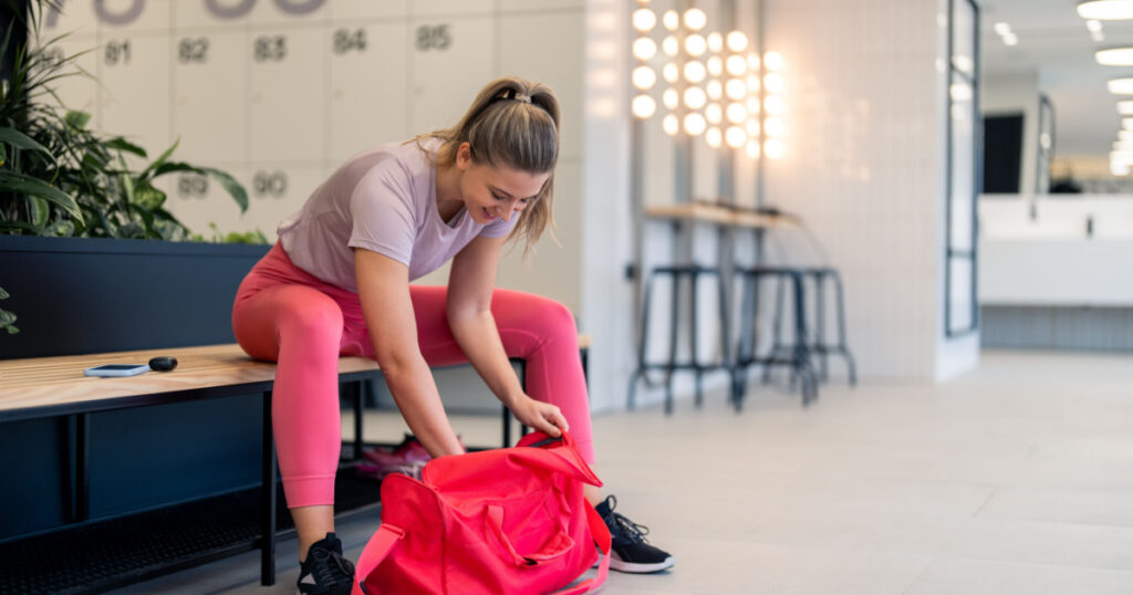 Beautiful satisfied athletic young woman in sportswear sitting on bench in locker room at gym, packing sports equipment in gym bag after finished workout fitness training session, preparing to go home