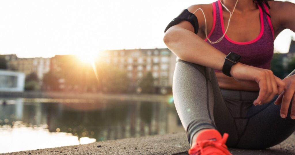 Young woman runner resting after workout session on sunny morning. Female fitness model sitting on street along pond in city. Female jogger taking a break from running workout.