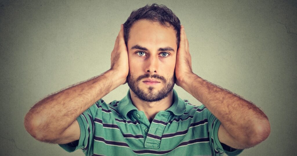 Closeup portrait handsome, peaceful, tranquil, looking relaxed, young man covering his ears, observing isolated on gray wall background. Hear no evil concept. Human emotion facial expression