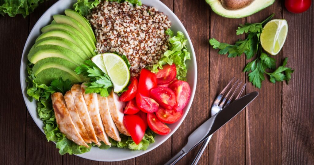 Healthy salad bowl with quinoa, tomatoes, chicken, avocado, lime and mixed greens, lettuce, parsley on wooden background top view. Food and health.