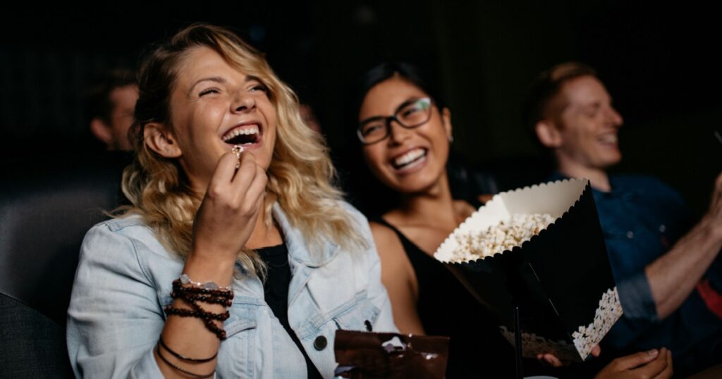 Young woman with friends watching movie in cinema and laughing. Group of people in theater with popcorns and drinks.