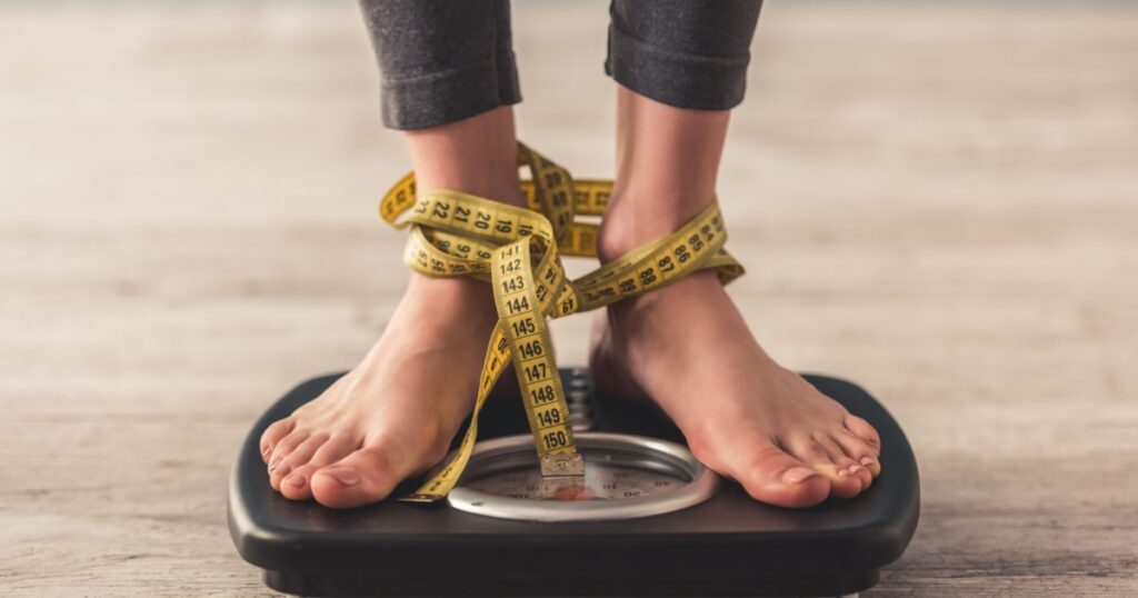 Cropped image of woman feet standing on weigh scales, on gray background. Legs winded with a tape measure