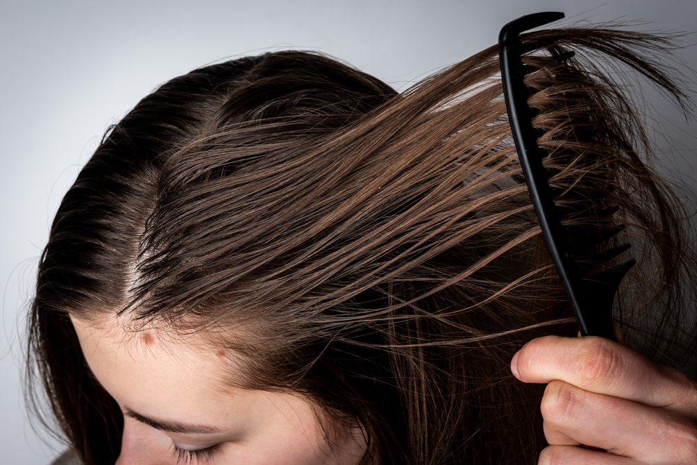Young woman combing dirty greasy hair on gray background.
