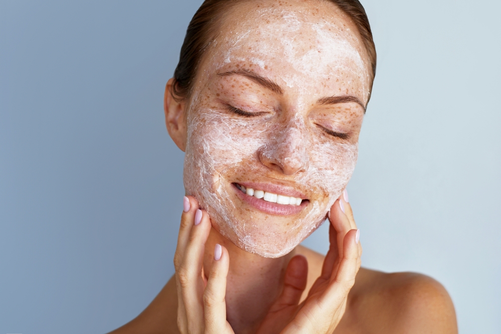 Self love. Skin care beauty portrait. Facial scrub, young happy woman with a freckles is applying white peeling cosmetic product to whole her face. Daily skincare routine. Dermatology. Exfoliating