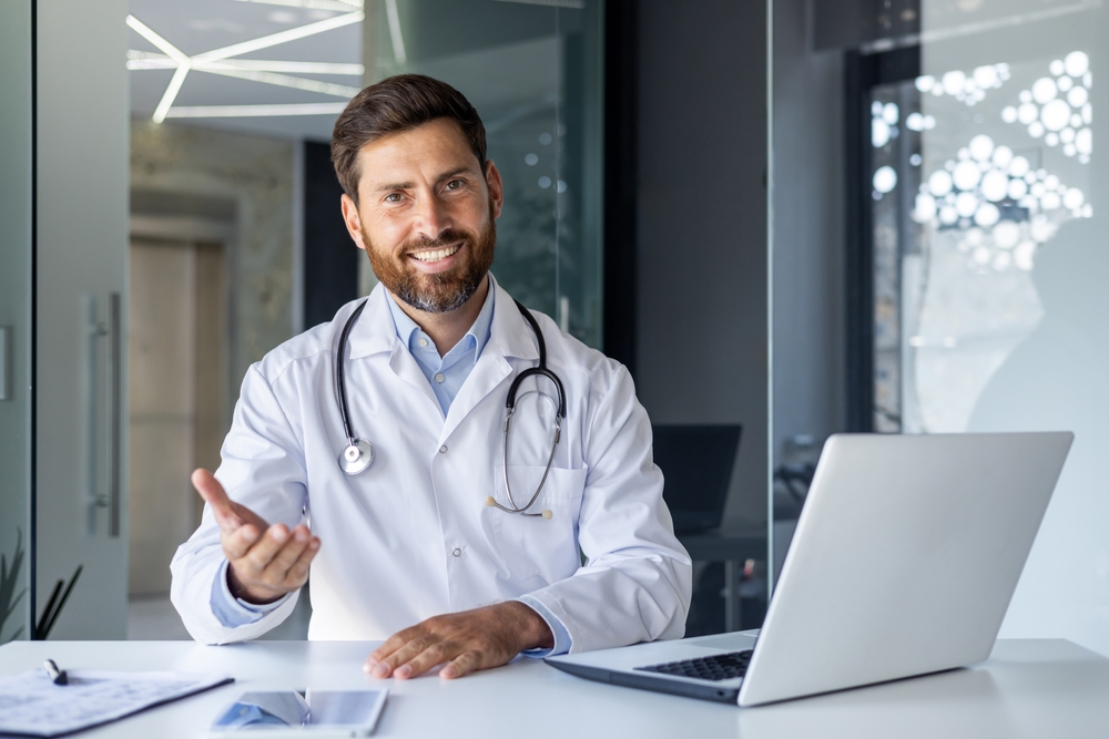 Portrait of a smiling young male doctor talking and consulting on a video call. He is sitting at the table in the office of the clinic, gesturing with his hand in front of the camera.