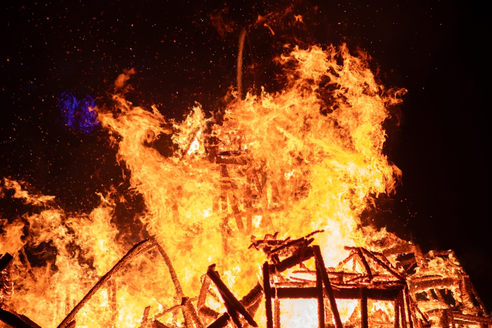 Dramatic scene of a house engulfed in intense flames in a dark night sky. The fire rages, engulfing the structure completely, creating a powerful image of destruction.