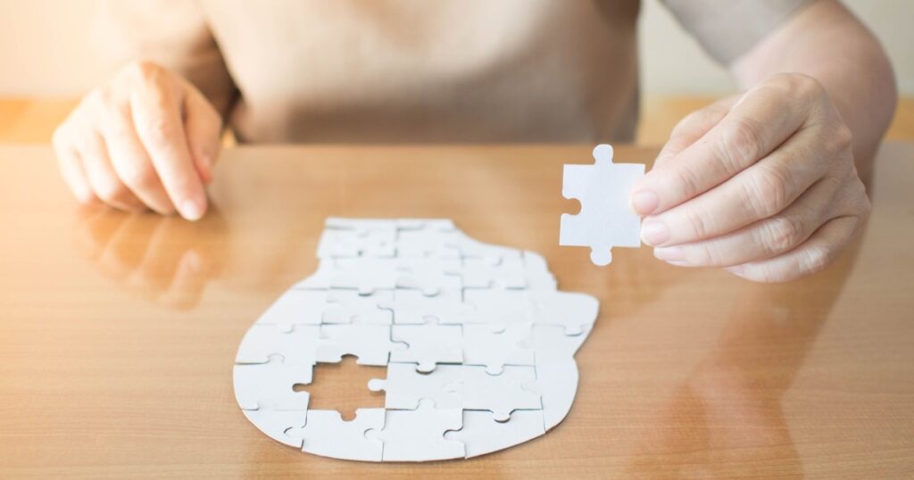 Elderly woman hands holding missing white jigsaw puzzle piece down into the place as a human head brain shape. Creative idea for memory loss, dementia, Alzheimer's disease and mental health concept.
