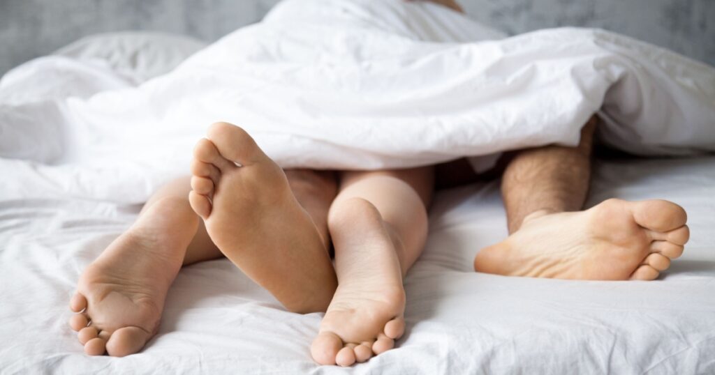 Close up of passionate lovers feet touch having sex on white sheet in bedroom, sensual couple enjoy intimate moment relaxing in bed, legs of boyfriend and girlfriend make love in woman on top position