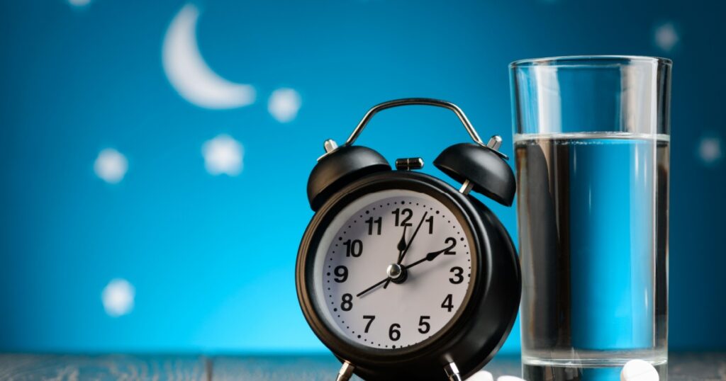 Alarm clock, pills and glass of water on a bedside table. Insomnia, treatment for sleepless nights.