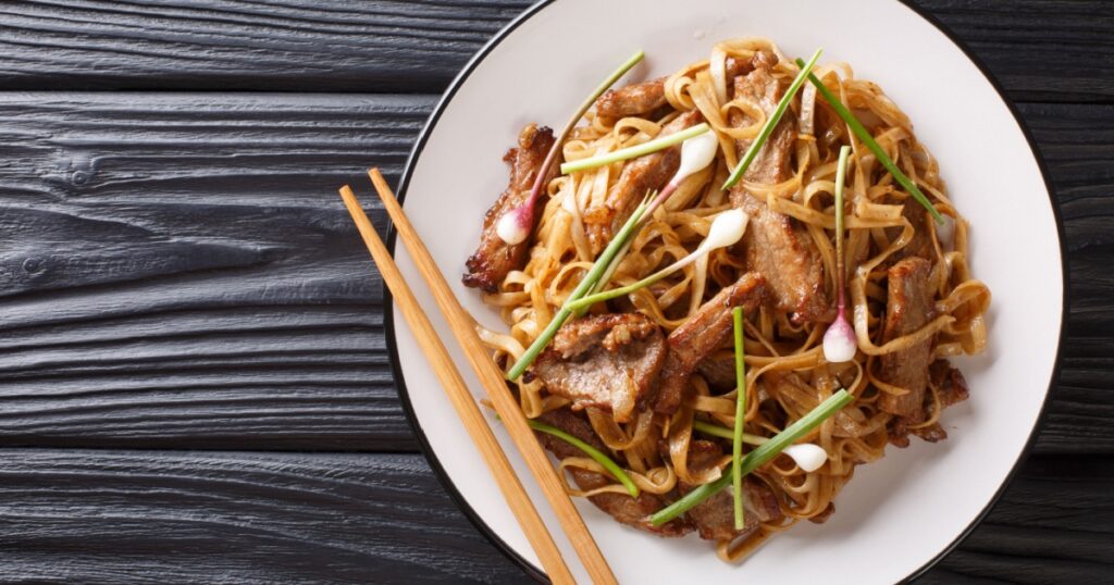 Beef Chow Fun a staple Cantonese dish, made from stir-frying beef, wide rice noodles and vegetables closeup on the plate on the wooden table. Horizontal top view from above