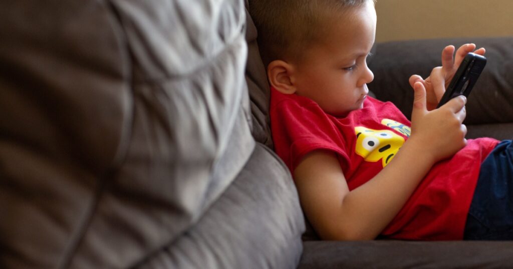 Charlotte, NC - July 1, 2019: Boy toddler using wireless device on a sofa with bad posture.