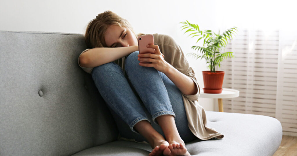 Portrait of beautiful young woman with depressed facial expression sitting on grey textile couch holding her phone. Cyber bullying victim concept. Sad female in her room. Background, copy space.