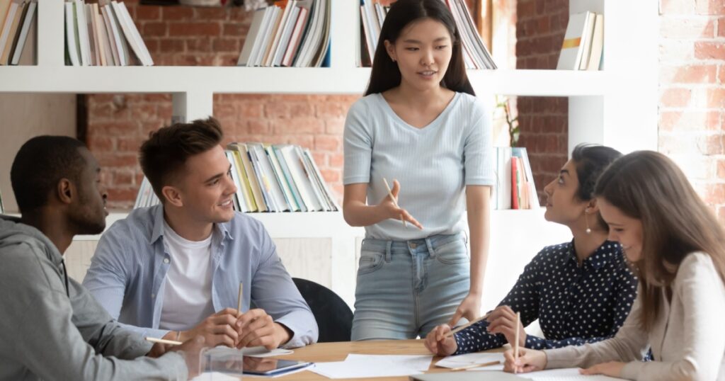 Smiling motivated asian female team leader holding meeting with diverse groupmates, explaining new project detail. African american, indian and caucasian concentrated students discussing assignment.