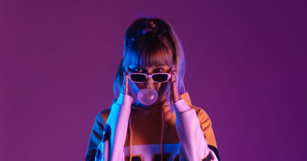 Pretty young 20s fashion teen girl model wear glasses blowing bubble gum looking at camera standing at purple studio background, igen teenager in trendy stylish night glow 80s 90s concept, portrait