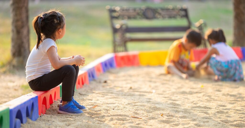 Little girl sitting lonely watching friends play at the playground.The feeling was overlooked by other people.Concept child shy.