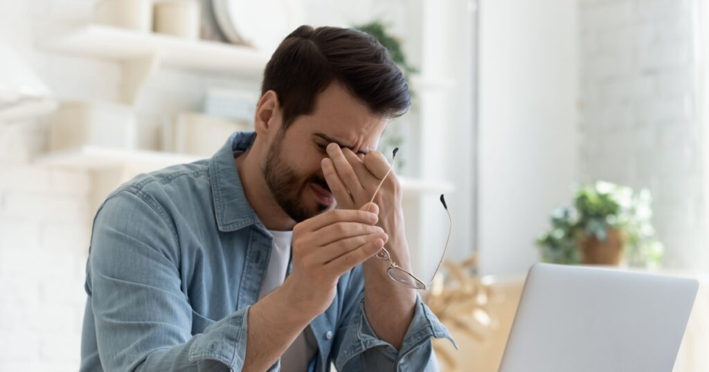 Tired young man feel pain eyestrain holding glasses rubbing dry irritated eyes fatigued from computer work, stressed man suffer from headache bad vision sight problem sit at home table using laptop