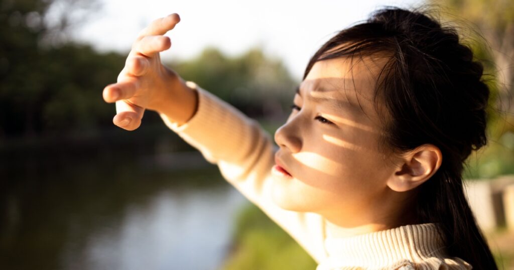 Asian child girl covering face by hand to protect,prevent face skin from bright sun in outdoor summer,female people hand covered the strong sun light feeling hot uncomfortable,risk of eyes damage