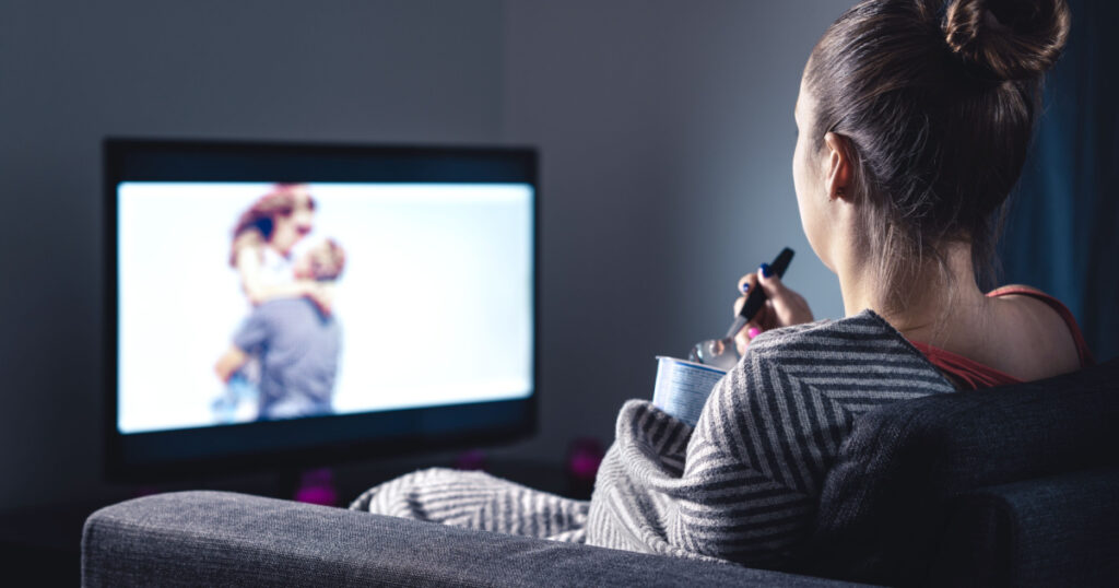 Woman watching romantic movie and eating ice cream. Sad lonely single girl streaming series or film on tv home at night. Person with stream on television. Comfy candid relaxing under blanket on couch.