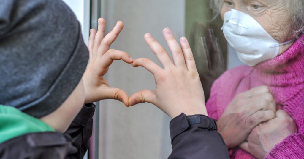 Grandmother mature woman in a respiratory mask communicates with her grandchild through a window. Elderly quarantined, isolated. Coronavirus covid-19. Caring with older people. Family values, love