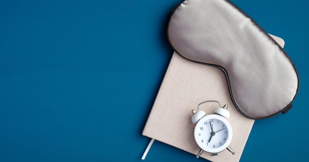 Flat lay composition with sleep eye mask, dream book and alarm clock on dark blue background. Healthy sleeping concept.