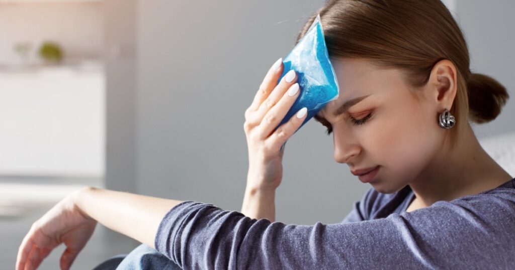 Woman applying ice pack as cold compress on forehead due to headache, migraine, tired after work, sitting indoors in apartment. Put cold on head for relief hurt and fever