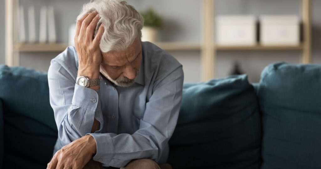 Frustrated unhappy middle aged mature man sitting on sofa, feeling depressed alone at home. Confused senior retired grandfather worrying about difficult life decision, copy space, old people solitude.