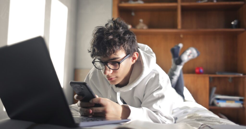 young boy studies lying on the bed using computer and smartphone
