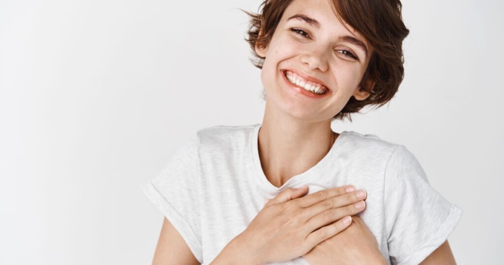 Close up of happy smiling woman say thank you, holding hands on heart grateful, express gratitude, standing against white background.