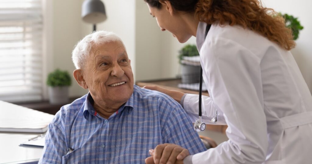 Close up caring nurse holding smiling mature patient hand at meeting in hospital, doctor caregiver wearing uniform comforting and supporting senior man, good news about treatment, empathy and care