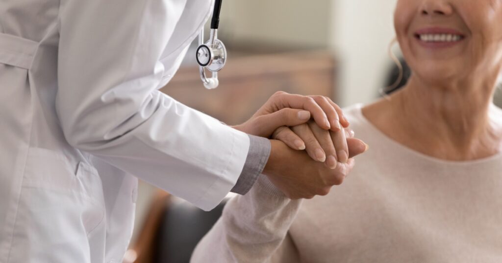 Doctor giving hope. Close up shot of young female physician leaning forward to smiling elderly lady patient holding her hand in palms. Woman caretaker in white coat supporting encouraging old person