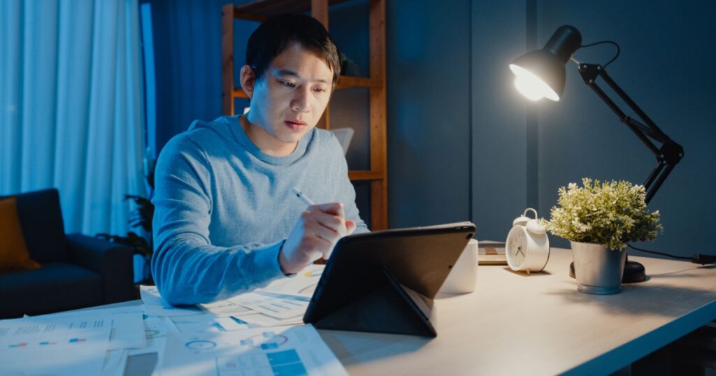 Asia freelance businessman focus work pen write on tablet computer busy with full of graph paperwork on desk in living room at home overtime at night, Work from home during COVID-19 pandemic