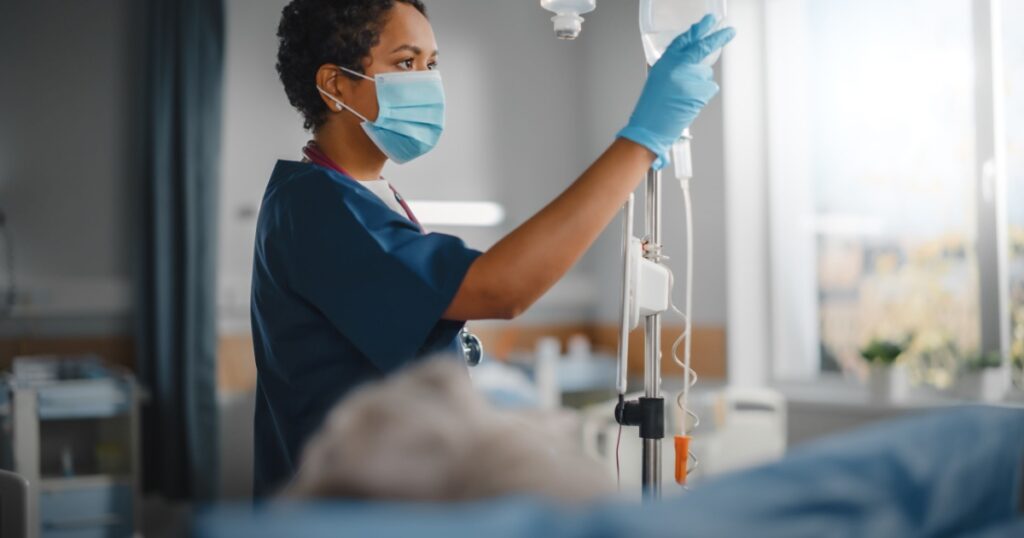 Hospital Ward: Professional Black Head Nurse Wearing Face Mask Does Checkup of Patient's Vitals, Checking Heart Rate Computer, Intravenous or Iv Fluids Drip Bag. Caring Nurse Monitors Person Recovery