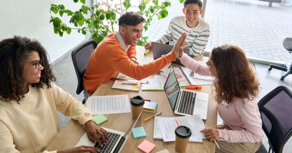 Young happy laughing creative team four multicultural coworkers or college students group celebrate success together giving highfive in office classroom. Top view. Great teamwork result high five
