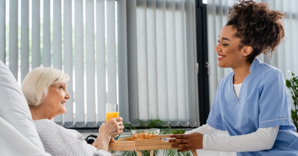 Side view of smiling african american nurse holding tray with food near patient with orange juice in hospital ward