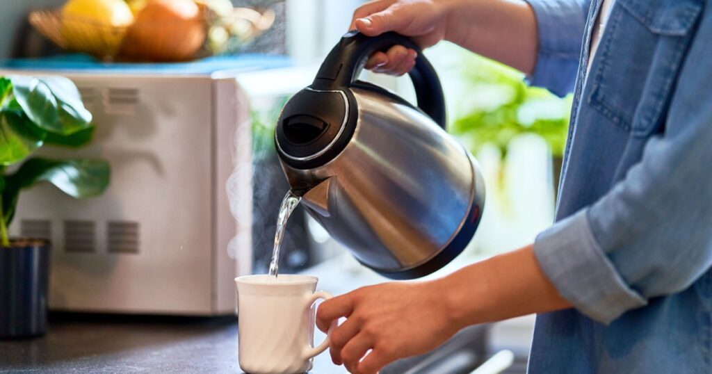 Female hands pouring boiling water from a modern metal stainless kettle in a glass cup for brewing tea in the kitchen at home