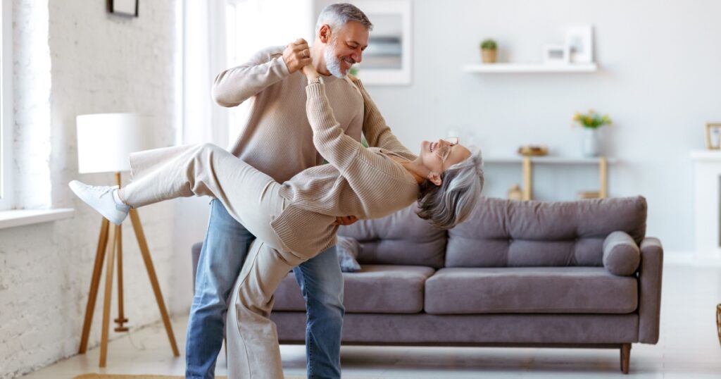 Keep moving. Romantic senior family couple wife and husband dancing to music together in living room, smiling laughing retired man and woman having fun, enjoying free time together at home