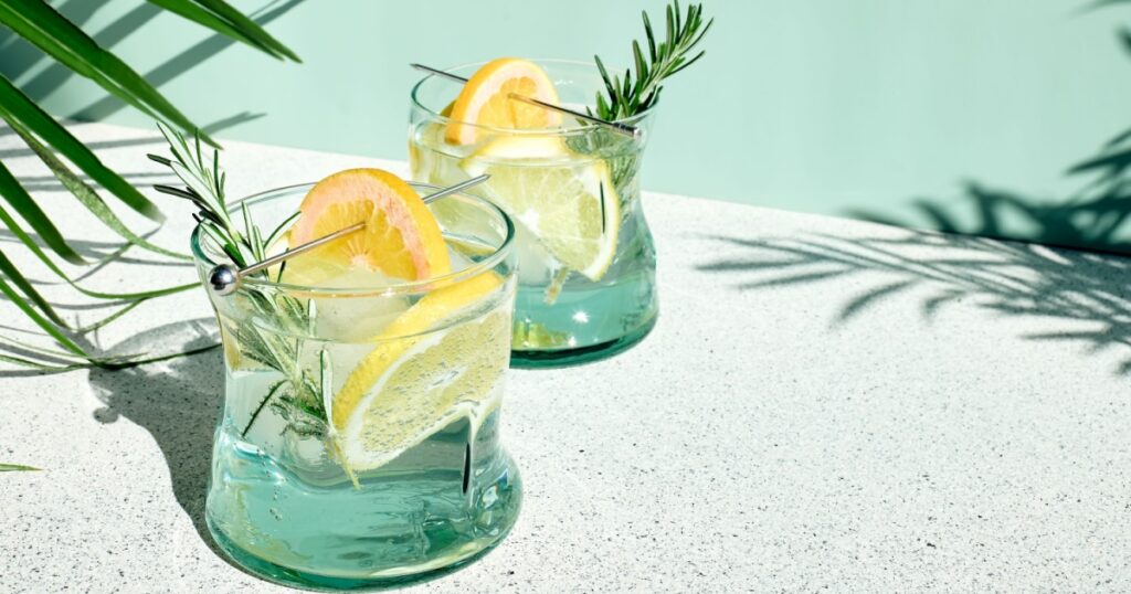 Summer refreshing lemonade drink or alcoholic cocktail with ice, rosemary and lemon slices on pastel light green surface. Fresh healthy cold lemon beverage. Water with lemon.