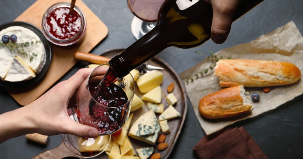 Man pouring red wine from bottle into glass over black table with snacks, top view