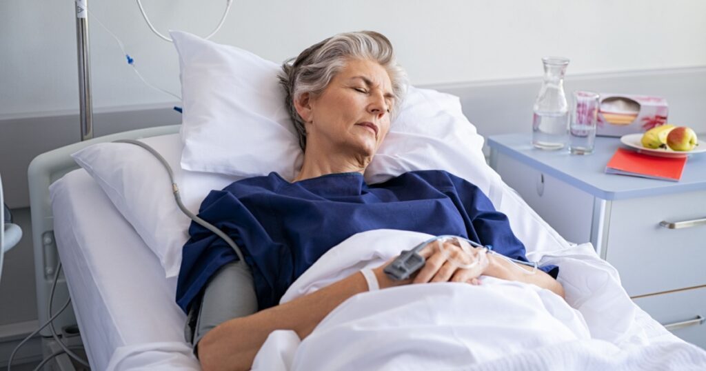 Elderly patient sleeping on a medical bed in hospital ward. Senior woman resting after operation with eyes closed and IV drip. Old mature woman lying on bed and wearing blue hospital gown.